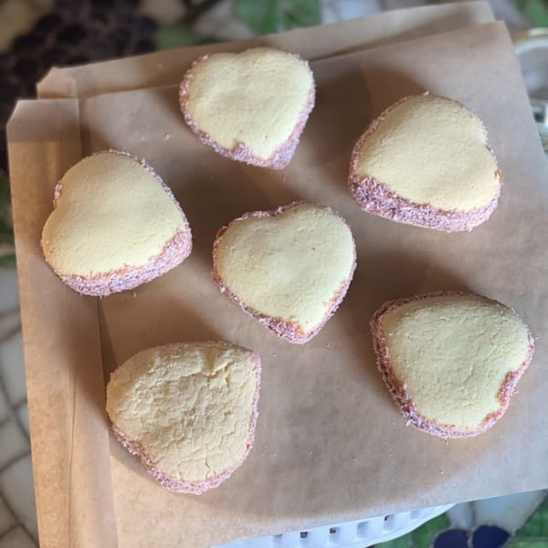 Six heart-shaped alfajores with pink sprinkles on the edges, arranged on brown parchment paper.