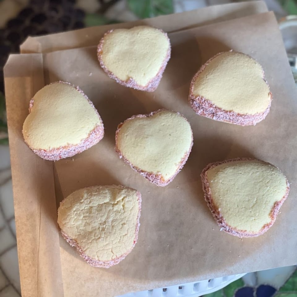 Six heart-shaped alfajores with pink sprinkles on the edges, arranged on brown parchment paper.